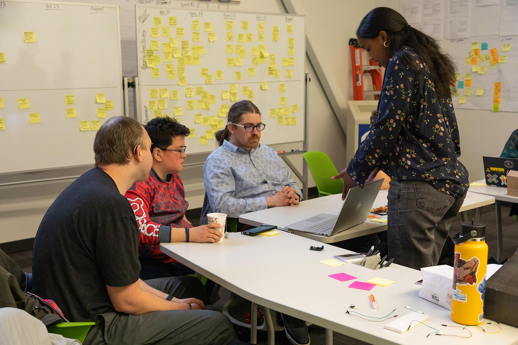 Woman showing laptop screen to three men with many workshop stickey notes on whiteboard behind