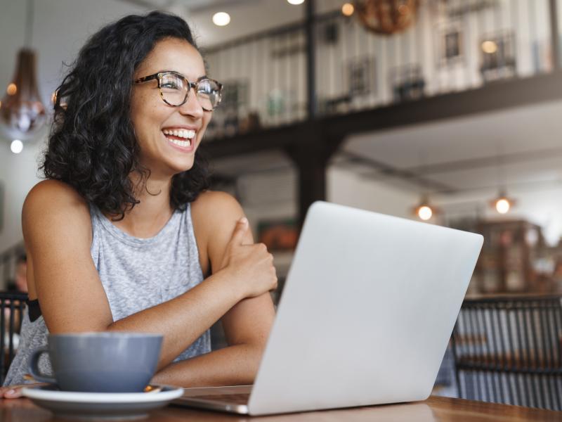 woman of color laughing and at table with laptop and coffee